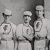 Three baseball players are standing together in their baseball uniforms. One player has his arms folded and another is holding a baseball.