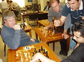A picture of four men looking over a chessboard and gesticulating. In the background are other chess boards on tables, set up differently.
