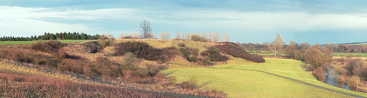 At left a grassyu hill with shrubbery, dropping to a meadow at centre. At right a narrow river winds by.