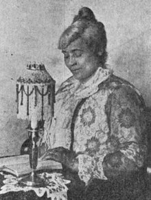 An older African-American woman with grey hair, seated at a table, hands and eyes on an open book; there is a small ornate lamp nearby, and lace on the tablecloth. She is wearing a lace or embroidered dress.