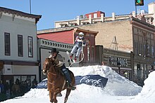 A horse and rider running down a snowy street, pulling a skier who is jumping an artificial hill made of snow