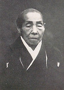 An older Japanese woman, wearing a kimono, greying hair dressed back to the nape