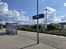 We can see a train platform featuring a sign indicating "Bremen Walle" as well as a waiting area with sitting a bit further back. Behind the tracks there is a bit of a residential area and the Bremen TV tower on the far left.