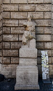 A damaged statue is on a pedestal in front of a stone wall; the pedestal has a number of pieces of paper with writing on them glued to it