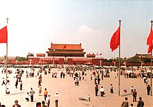 A large crowd of people gather in an open space with multi-level red buildings in the background, and three flag poles with red flags in the foreground.