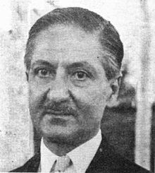 A middle-aged man wearing a suit. He has a mustache and slicked-back hair and is looking just to the camera's left.