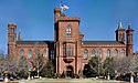 A brick building, reminiscent of a castle, slightly obscured by bare trees. There is a large central tower with the entrance at its base and a smaller tower at each corner of the building, each with a varying design.