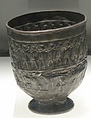 A silver cup with two rows of engraving. On top 9 human figures bearing cups or vases, below them three deer a hind with head aloft follows and is followed by stags with lowered heads