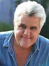 A gray-haired man with a prominent chin wearing a blue shirt and smiling