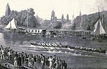 A river scene, with two eight-oared boats racing in the middle of the river, one just in front of the other. There are crowds on each bank and some sailing boats and barges by the far bank. In the distance, trees and church spires.