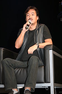 一名坐在黑色皮椅上，手持麦克风，身着黑衬衫、黑皮革背心、黑牛仔裤的男人