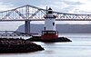 A red, white and black lighthouse at dusk with a large bridge in the background