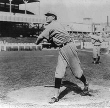 A man wearing a baseball uniform stands truned–his right having just pitched a baseball from the pitcher's mound.