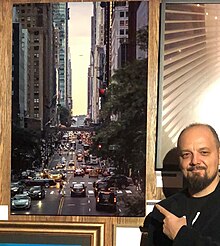 Burak Karavit stands in front of his work in the Dean Collection. Pointing to his work with his finger, a street of New York appears in Karavit's work.