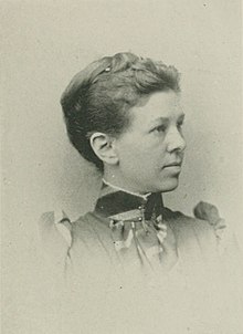 B&W portrait photo of a woman with her hair in an up-do, wearing a high-collared, dark blouse.