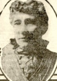 An older white woman with curly grey hair in an updo, wearing a high-collared ruffled blouse