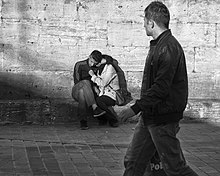 A man on a brick street walking by and looking on another man and woman sitting and embracing each other in the background