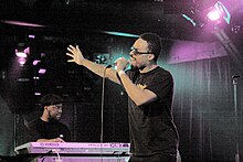 A man dressed in t-shirt, jeans, and sunglasses, singing into a microphone, accompanied by a similarly dressed man on keyboards in the background, on a dimly-lit stage
