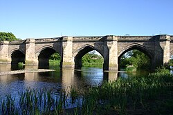 Four arches of a stone bridge straddling a slow-flowing river, with green banks on eithers side