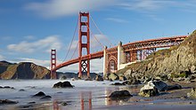 The Golden Gate Bridge, a two-tower suspension bridge painted a dull orange, seen on a sunny day from a beach with mist over the water.