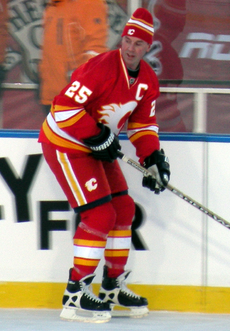 A hockey player in a red and white uniform skates up the ice in anticipation of receiving a pass. He is wearing a toque instead of a helmet