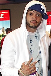 A shot of Travie McCoy, holding a piece sign with his fingers into a nearby camera.