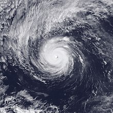 A satellite image of a powerful hurricane over the Eastern Pacific Ocean, with a cloudy but well-defined eye, a central region of deep convection marked by thick clouds, and several spiral bands; a large arc of thin high clouds is fanning out to the northwest of the hurricane