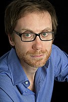 A blue-eyed, ginger-bearded man in a blue shirt and glasses looking into the camera before a black background.