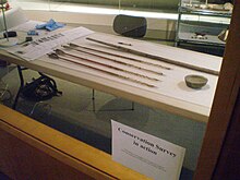 Behind a pane of glass, four American Indian arrows rest on a plastic card table, surrounded by scientific equipment. A printed sign on the window states "Conservation Survey in action".