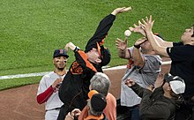 A group of men mostly wearing orange and black baseball caps and shirts in various configurations stand at their seats near the edge of a baseball field, leaning backwards slightly with their hands stretched with a baseball in the air between them. From the field a player in the Boston Red Sox road uniform looks on.