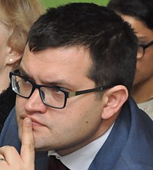 close-up of Jan Chvojka wearing glasses and holding his left index finger up to his chin, surrounded by other people