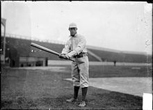 A man in a white baseball uniform with dark socks poses with his baseball bat extended.