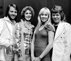 Two long-haired men, one with a beard, and two women, one with dark curly hair and one with blonde hair, standing together and all smiling