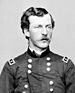 Head and shoulders of a young white man with wavy hair and a mustache, wearing a double-breasted military jacket with a rectangular patch over each shoulder.