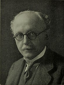 Black-and-white portrait of a bespectacled, balding man with a moustache