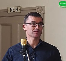 Man with glasses speaking at a microphone