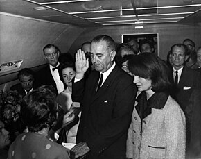 Lyndon B. Johnson raises his hand above an outstretched Bible as he is sworn in as President as Air Force One prepares to depart Love Field in Dallas. Jacqueline Kennedy, still in her blood-spattered clothes (not visible), looks on.