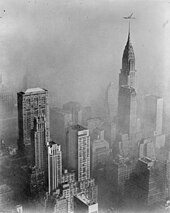 Photograph of skyscrapers seen from a great height, surrounded by smog. Unlike the previous photo of the 1966 smog, no horizon can be seen as the entire sky is blotted out by the smog. If the prior photo's vantage point seems to be "above" a blanket of smog, this photo is completely underneath and within it.