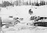 View of Stapp Lakes Lodge with snow on the ground. Log cabins, two small four-wheeled wagons and a truck visible beneath a ridge