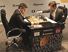 Ian Nepomniachtchi and Magnus Carlsen both sitting down at a chessboard during a game. Both men are wearing suits and ties. Both the table they are sitting at and the walls around them bear numerous logos of the event sponsors. Nepomniachtchi has the White pieces, and Carlsen has the Black pieces.