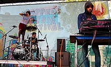 Crystal Castles performing on stage at a music festival. A drummer is seated behind a Yamaha drum set, and Alice Glass is standing on the drum set, singing into a microphone. Ethan Kath is on the right, operating an electronic keyboard while wearing a hooded sweatshirt.