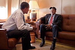 Photograph of Mays in a suit and a baseball cap sitting on a sofa facing President Barack Obama waering a dress shirt and suit pants; airplane windows are visible to their left