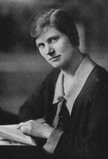 A white woman with side-parted straight dark hair, wearing a collared dress with one hand on a book