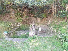 Photograph of a Ryukyuan well at the bottom of a cliff. The well presents several pools on the left side, all enclosed with stone walls, and a paved area on the right side, separated from the pools by a 80 cm high stone wall. The space in front of the pools and paved area is paved as well, but more roughly and grasses grow through the stones of the pavement. The cliff in the background is covered in vegetation as well. There is an incense burner on the left side of the pools, at the bottom of the cliff.