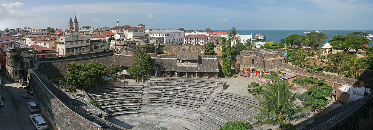 A panoramic photograph with an amphitheater surrounded by a castle wall in center. The dull colors are interrupted by green trees and beyond the walls are white houses with red tiled roofs and a blue ocean.