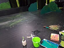 containers and trays of coloured paint, an black floor, paint brushes, a splash of water, and some scenic elements around the edge of the floor