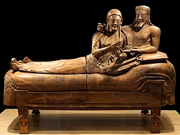 A sculpture of a woman and man reclining together on a couch, their upper bodies to the right and their legs to the left. There is a marked contrast between the high relief busts of their upper bodies and the very flattened lower bodies and legs. They have almond-shaped eyes and long braided hair, and are smiling widely. The man has a beard that is bobbed. The woman's hands are gesticulating in front of her as if she was holding something that is no longer there, or perhaps gesturing while speaking. The man has his right arm draped around the woman's shoulders in an intimate pose, and his right hand on her shoulder also appears to once have held some item. His left hand rests palm up in the crook of the woman's left elbow.