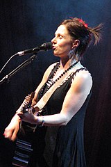 She is shown in upper-body shot and left profile. She has her eyes closed as she sings into a microphone and plays her guitar.