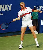 A blonde, bearded man in a white and red polo shirt swings his arm during a tennis rally