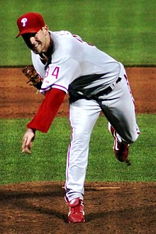 A man in a gray baseball uniform with red trim and the number "34" on the sleeve in red and a red baseball cap throwing a baseball (unseen) from a dirt mound